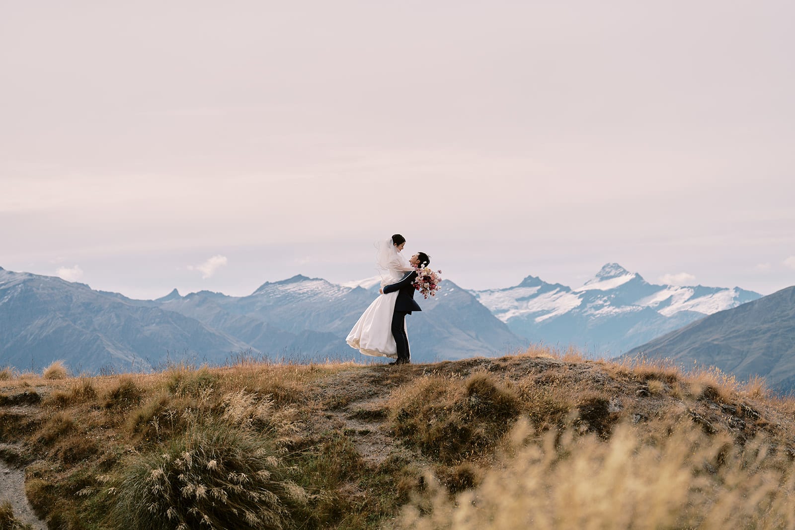 Queenstown Tekapo New Zealand Heli Wedding Elopement Pre-Wedding Shoot Photographer クイーンズタウン　テカポ　ニュージーランド　エロープメント 前撮り　フォトウェディング　結婚式 | A couple in wedding attire embracing on a hill with mountain scenery in the background, captured seamlessly by a renowned Queenstown Wedding Photographer.